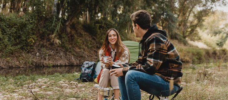 na imagem, vemos um casal conversando em um área aberta. ao fundo, uma barraca de camping, fazendo referência aos tipos de hospedagem existentes no artigo.