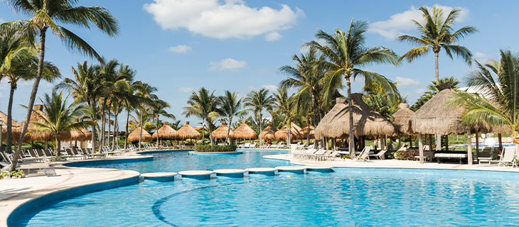 na imagem, vemos uma área de piscina e alguns bangalôs e cadeiras espriguicadeiras, como vemos em um resort.