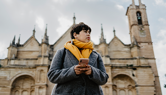 na imagem, vemos um turista segurando um celular, em frente a uma igreja, simbolizando o turismo religioso.