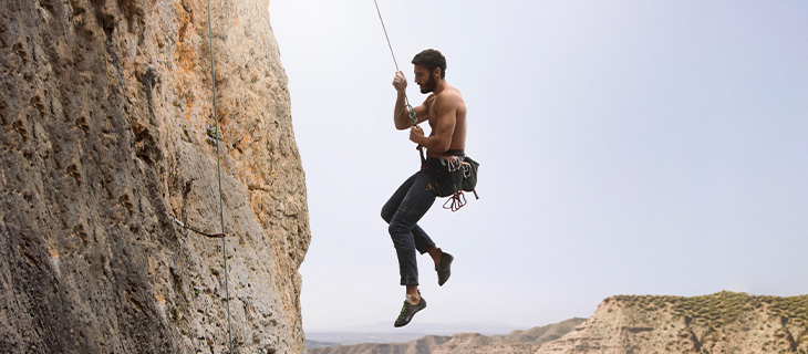 na imagem, vemos um homem sem camisa, de calça jeans e sapatos, segurando por um cabo de aço enquanto desce uma rocha, praticando o esporte rapel.